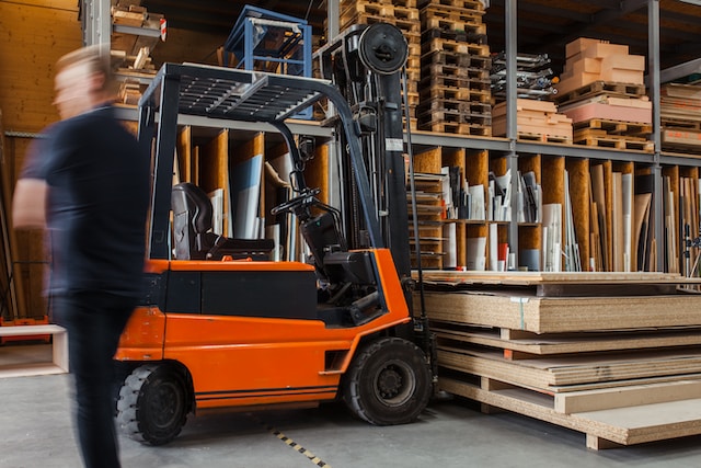 photo in wood warehouse with a forklift in the middle of the photo and a blurry person to the left of the photo.